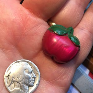May include: A red and green plastic apple button with a shiny finish. The button is about the size of a US nickel.