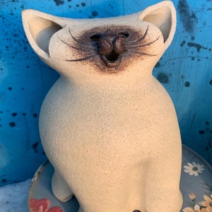 May include: A ceramic cat figurine with a smiling face and large ears. The cat is off-white with brown accents on the face and whiskers. The figurine is sitting on a floral patterned surface against a blue background.