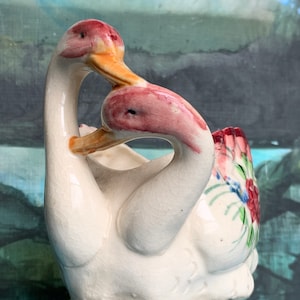 May include: A pair of white ceramic swans with pink and yellow accents. The swans are intertwined with their necks touching. The swan on the right has a floral design on its back.