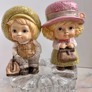 May include: A pair of ceramic figurines of children, a boy and a girl, wearing hats and carrying suitcases. The boy is wearing a brown and green plaid shirt and brown pants. The girl is wearing a pink hat and a green dress. They are standing on a clear glass base.