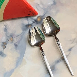 May include: A set of two silver salad serving utensils with white handles. The utensils are on a white and blue marble surface with a red watermelon slice prop.