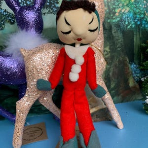 May include: A handmade doll in a red jumpsuit with a white collar and a white pom-pom. The doll has a fabric face with closed eyes and dark hair. Teal gloves and boots complete the outfit. Glittery deer figurines are in the background.