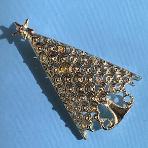 May include: A gold-toned Christmas tree brooch with a star on top. The brooch has an openwork design with iridescent accents. The tree is set against a light blue background.