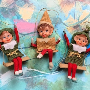 May include: Three vintage Christmas ornaments of elves on swings. The elves are wearing green and red outfits with gold trim. They have brown hair and smiling faces.