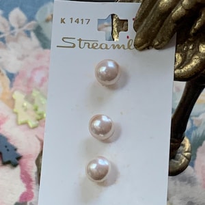 May include: Three pale pink pearl buttons on a white card. The card has the text "Streamline" and the number "K 1417" printed on it. The buttons are round and have a glossy finish. The background is a floral pattern.