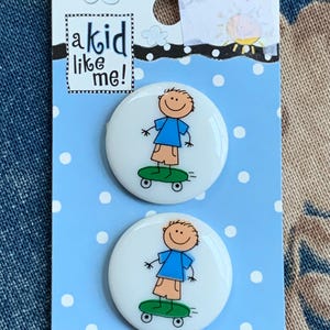 May include: Two round white buttons featuring a cartoon skater design, presented on a light blue card with white polka dots. The card reads "a kid like me!" and has a price tag of £1.25.