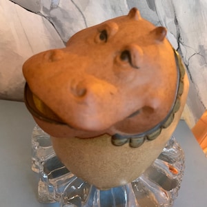 May include: A ceramic hippo figurine with a light brown head and a tan body. The hippo has small ears and a detailed face. The figurine sits on a clear, glass, flower-shaped base. The hippo has a decorative collar.