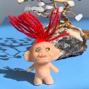 May include: A small plastic troll doll with red yarn hair and green glitter eyes. The troll is standing on a blue surface.