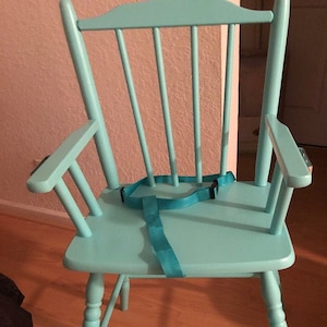 Puede incluir: Una silla de madera de color turquesa claro con un respaldo alto de barrotes verticales, dos reposabrazos y un asiento plano. Una correa de seguridad de nailon turquesa a juego con una hebilla de pl&aacute;stico negra est&aacute; sujeta al asiento.