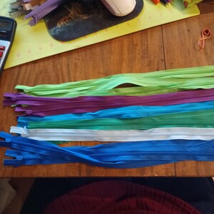 May include: A collection of colorful zippers in various shades of green, purple, blue, and white, arranged on a wooden surface. These zippers are ideal for sewing projects and crafts.