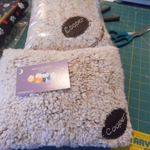 May include: Two beige, fluffy pet beds with a brown patch that says "Cooper." One bed is in a clear plastic bag. A business card with owl illustrations is on top of the other bed. The background is a green cutting mat.