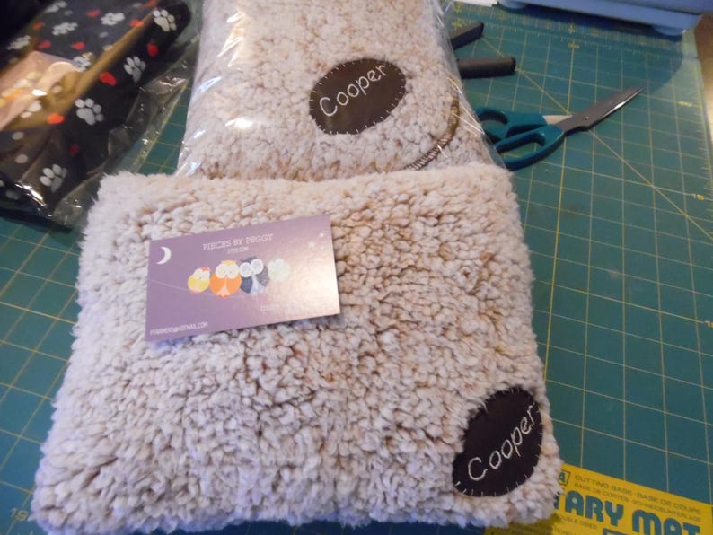 May include: Two beige, fluffy pet beds with a brown patch that says "Cooper." One bed is in a clear plastic bag. A business card with owl illustrations is on top of the other bed. The background is a green cutting mat.