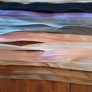 May include: Assortment of colorful zippers in various shades, including pink, beige, black, and lavender. The zippers are arranged horizontally, showcasing their different colors and textures. The image is taken from a close-up perspective, highlighting the details of the zipper tape and teeth.