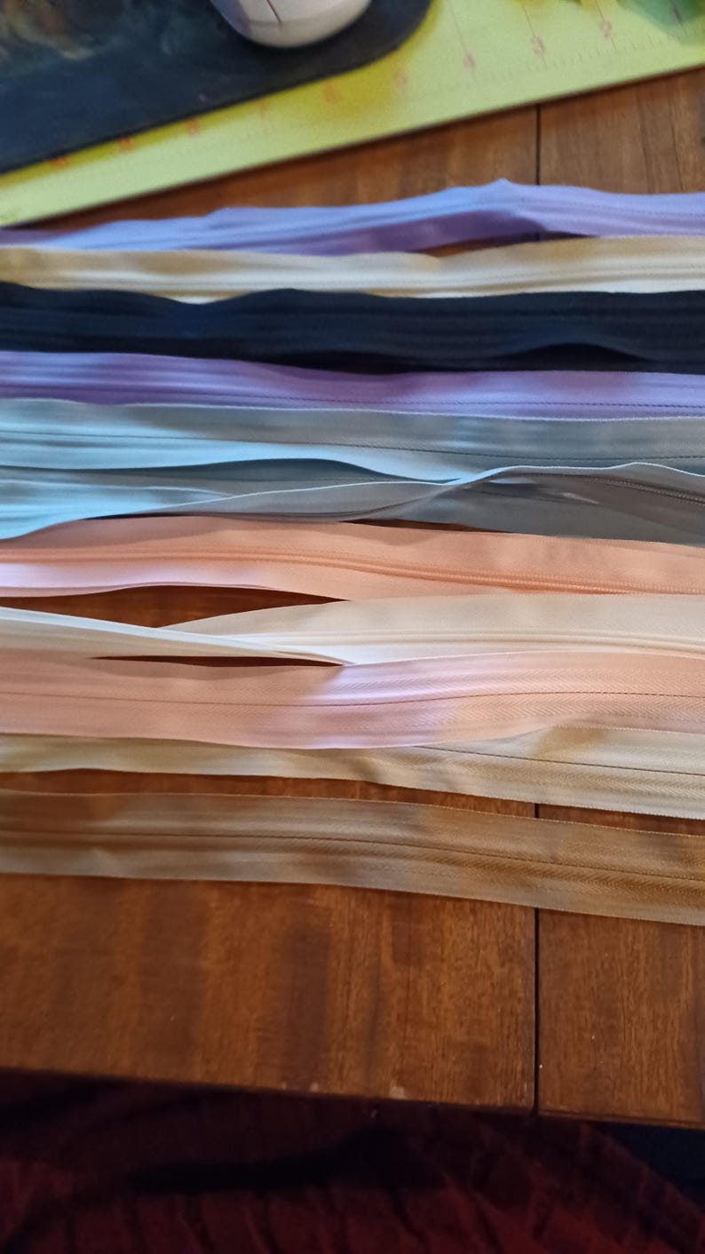 May include: Assortment of colorful zippers in various shades, including pink, beige, black, and lavender. The zippers are arranged horizontally, showcasing their different colors and textures. The image is taken from a close-up perspective, highlighting the details of the zipper tape and teeth.