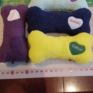 May include: A collection of plush dog bone-shaped toys in various colors: purple, light blue, navy, and yellow. Each bone has a heart-shaped patch with a pet's name: "Daphne", "Skeeter", "Wallaby", and "Gator". The toys are made of a soft, textured material.