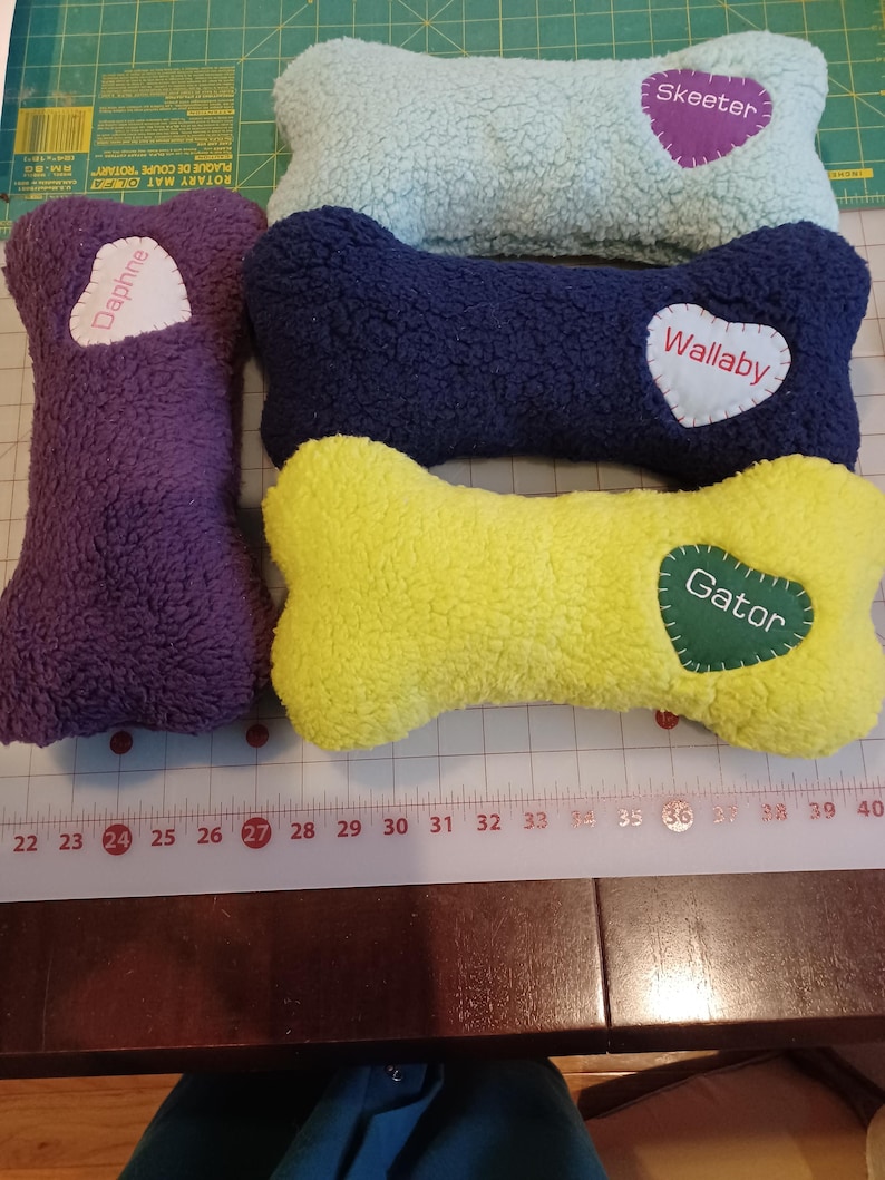 May include: A collection of plush dog bone-shaped toys in various colors: purple, light blue, navy, and yellow. Each bone has a heart-shaped patch with a pet's name: "Daphne", "Skeeter", "Wallaby", and "Gator". The toys are made of a soft, textured material.