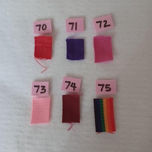 Puede incluir: Seis muestras de tela rectangulares dispuestas en dos filas, cada una etiquetada con una tarjeta de papel rosa numerada del 70 al 75. Las muestras incluyen rojo s&oacute;lido, morado, rosa, rosa claro, rojo oscuro y un patr&oacute;n de rayas arco&iacute;ris.