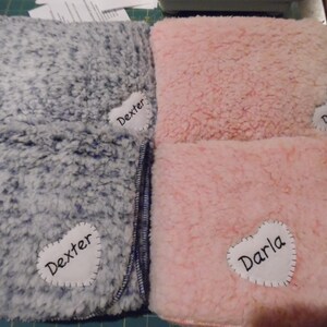 May include: Two plush blankets, one gray and one pink, are folded and displayed. Each blanket has a white heart-shaped patch with a name stitched on it: "Dexter" on the gray blanket and "Darla" on the pink one. The blankets have a soft, textured appearance.