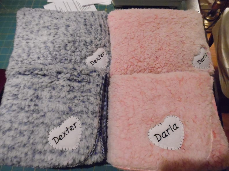 May include: Two plush blankets, one gray and one pink, are folded and displayed. Each blanket has a white heart-shaped patch with a name stitched on it: "Dexter" on the gray blanket and "Darla" on the pink one. The blankets have a soft, textured appearance.