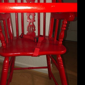 Puede incluir: Una trona de madera de color rojo brillante con un dise&ntilde;o de respaldo de husillos con un recorte decorativo central, una barra de seguridad horizontal y una correa de sujeci&oacute;n de tela roja con una hebilla de pl&aacute;stico negro.