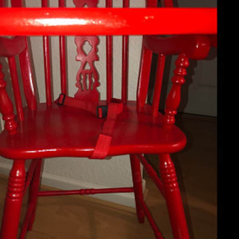 Puede incluir: Una trona de madera de color rojo brillante con un dise&ntilde;o de respaldo de husillos con un recorte decorativo central, una barra de seguridad horizontal y una correa de sujeci&oacute;n de tela roja con una hebilla de pl&aacute;stico negro.