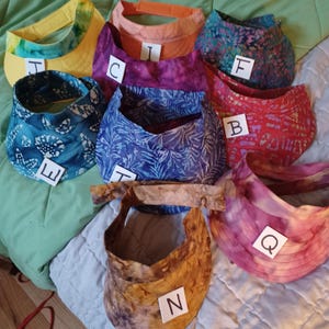 May include: A collection of nine fabric sun visors in various colors and patterns, including yellow, orange, purple, blue, red, and brown, each labeled with a white tag featuring a single letter from J, I, F, C, B, E, H, N, and Q.