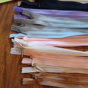 May include: A collection of various colored zippers, including black, beige, purple, gray, and peach. The zippers are laid out on a wooden surface, showcasing their different shades and styles. Ideal for sewing and crafting projects.