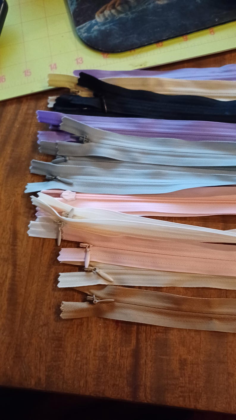 May include: A collection of various colored zippers, including black, beige, purple, gray, and peach. The zippers are laid out on a wooden surface, showcasing their different shades and styles. Ideal for sewing and crafting projects.