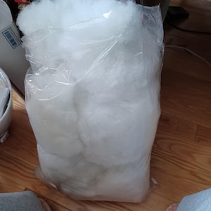 May include: A large clear plastic bag filled with fluffy white polyester fiberfill. The bag is partially transparent, revealing the soft, cloud-like texture of the filling. The bag is sitting on a wooden floor.