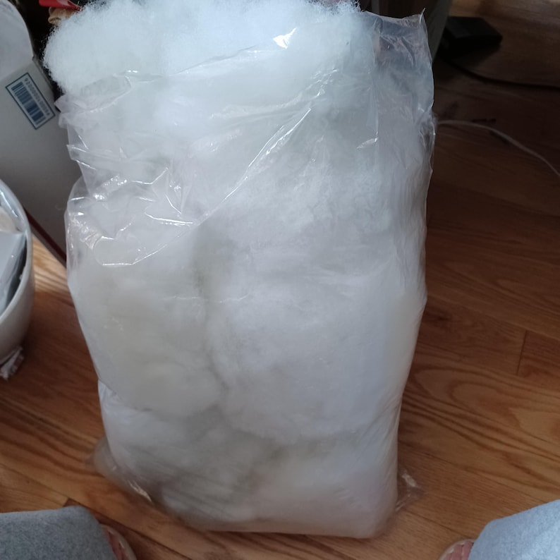 May include: A large clear plastic bag filled with fluffy white polyester fiberfill. The bag is partially transparent, revealing the soft, cloud-like texture of the filling. The bag is sitting on a wooden floor.