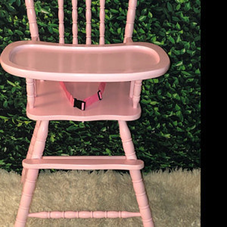 Puede incluir: Una trona de madera de color rosa claro con respaldo de husillos, una bandeja ancha, un reposapi&eacute;s y una correa de seguridad rosa con una hebilla de pl&aacute;stico negra. La silla tiene un acabado pintado liso y un dise&ntilde;o cl&aacute;sico con patas torneadas.