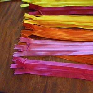 May include: A collection of colorful zippers in shades of yellow, burgundy, orange, pink, and fuchsia. The zippers are arranged on a wooden surface, showcasing their vibrant hues and design. Ideal for sewing and crafting projects.