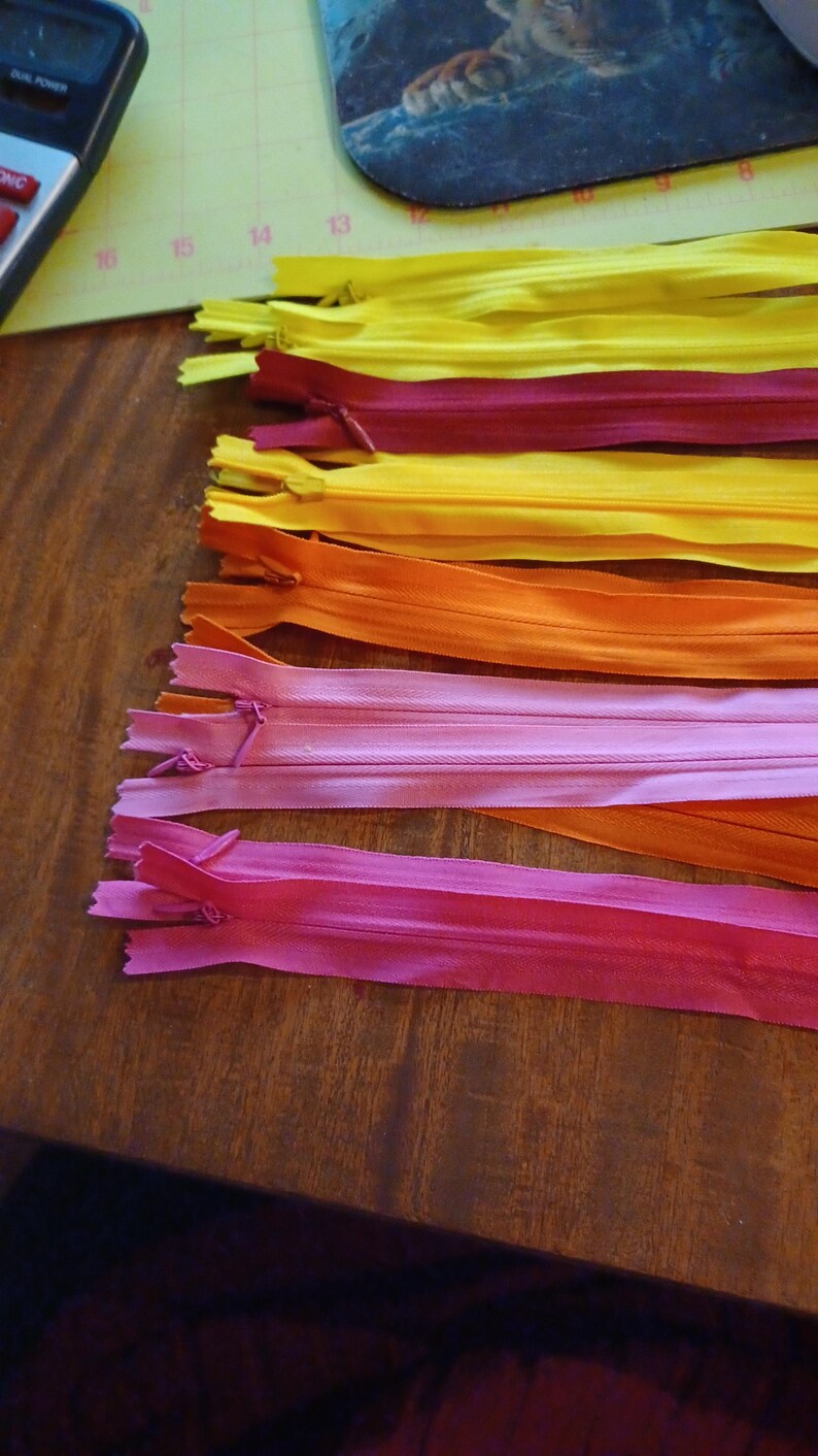 May include: A collection of colorful zippers in shades of yellow, burgundy, orange, pink, and fuchsia. The zippers are arranged on a wooden surface, showcasing their vibrant hues and design. Ideal for sewing and crafting projects.