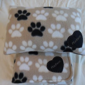 May include: A tan fleece pillow and blanket set featuring a repeating pattern of black and white paw prints. Each item has a black heart-shaped patch with the name "Madison" embroidered in white.
