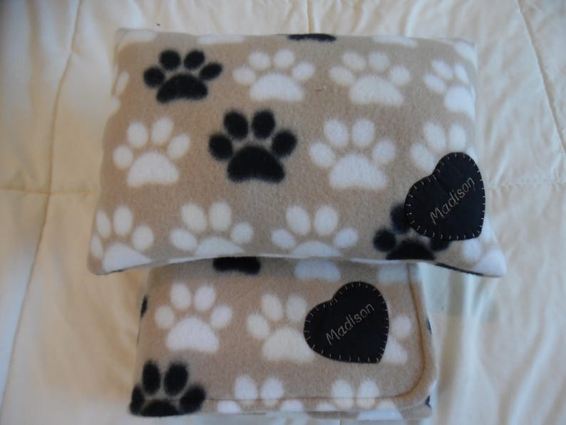 May include: A tan fleece pillow and blanket set featuring a repeating pattern of black and white paw prints. Each item has a black heart-shaped patch with the name "Madison" embroidered in white.