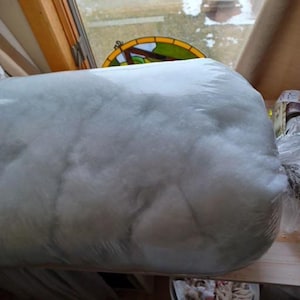May include: A large, cylindrical package of white polyester fiberfill, tightly wrapped in clear plastic. The fill appears fluffy and soft, suitable for stuffing pillows or crafts. The package is resting on a wooden surface.