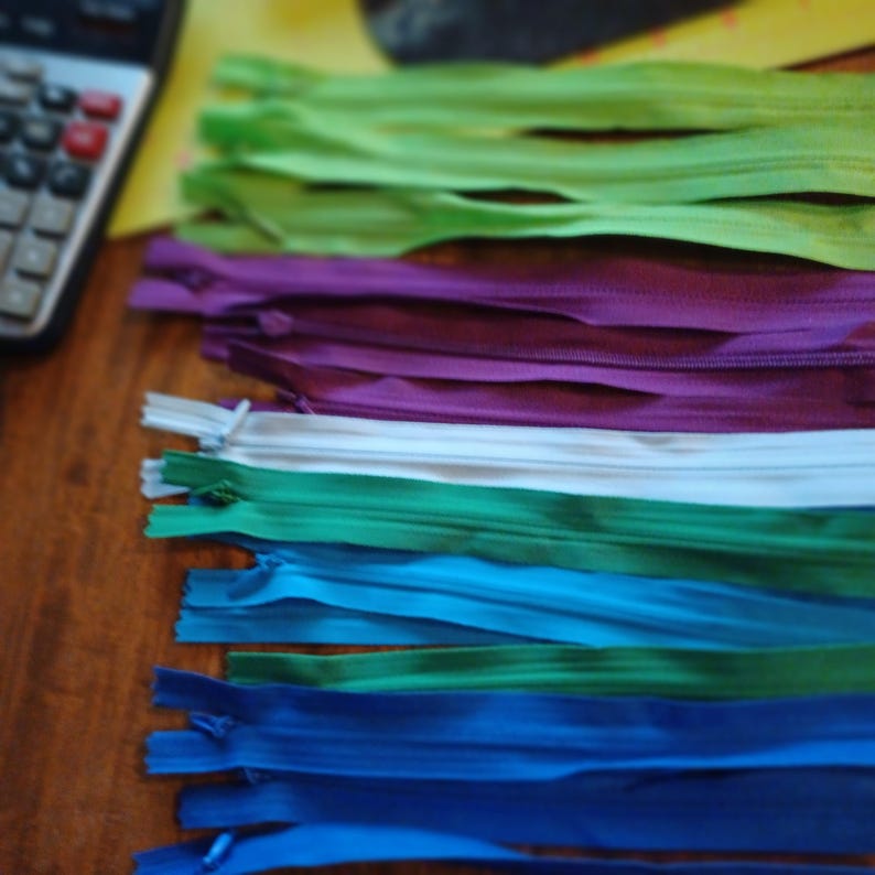 May include: A collection of colorful zippers in various shades, including green, purple, white, teal, blue, and navy. The zippers are arranged in a row, showcasing their different colors and textures. A calculator is visible in the background.