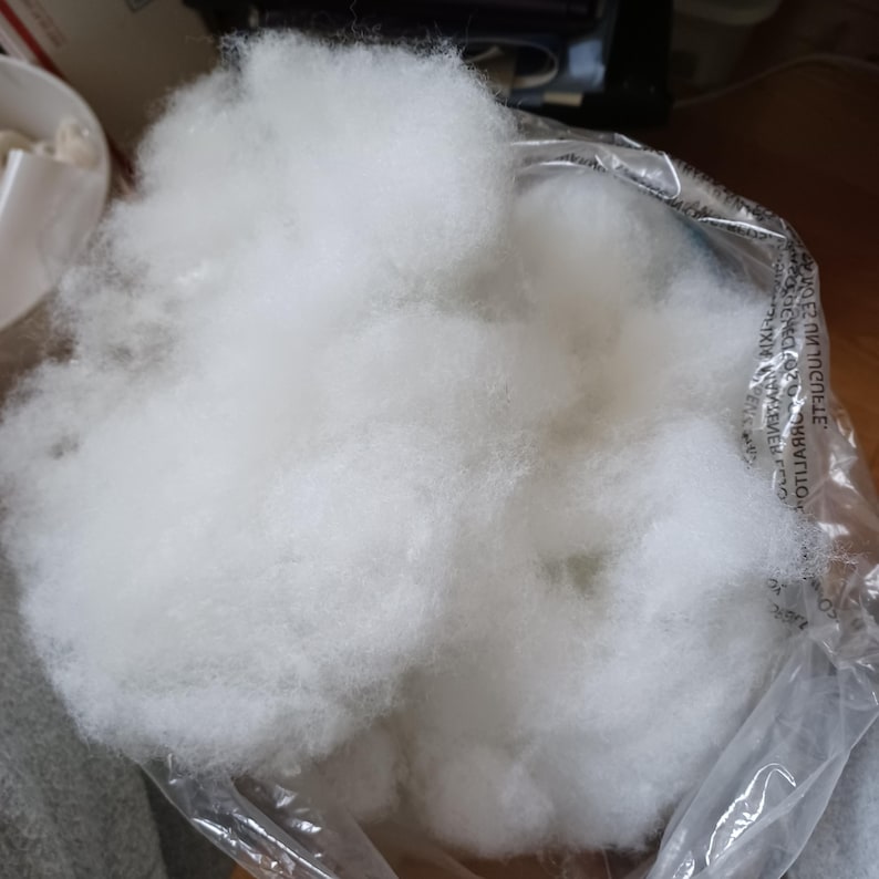 May include: A clear plastic bag filled with fluffy, white polyester fiberfill. The fiberfill is soft and cloud-like, perfect for stuffing pillows, cushions, or craft projects. The bag is partially open, revealing the contents.