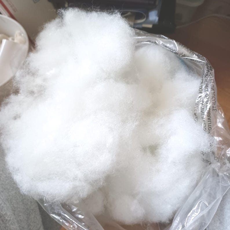 May include: A clear plastic bag filled with fluffy, white polyester fiberfill. The fiberfill is soft and cloud-like, perfect for stuffing pillows, cushions, or craft projects. The bag is partially open, revealing the contents.