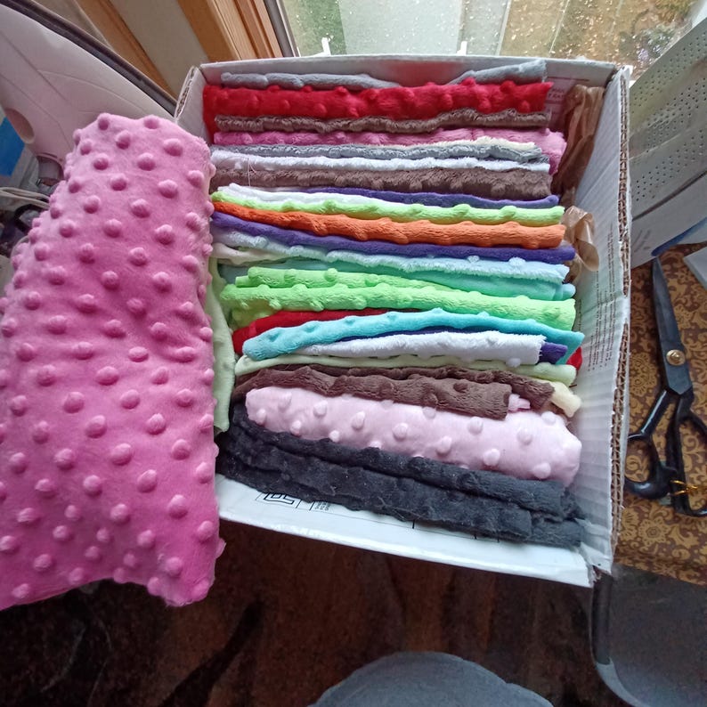 May include: A box filled with various colors of dimple dot fabric. A large roll of pink dimple dot fabric is on the left. The box contains folded fabric in red, gray, orange, green, blue, brown, and pink.