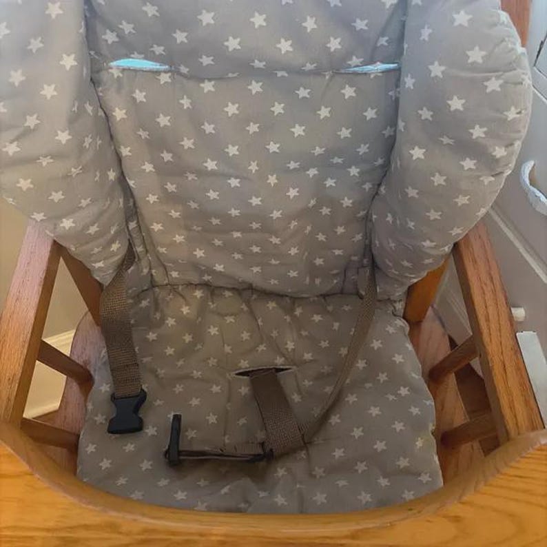 Puede incluir: Un coj&iacute;n para trona de tela gris claro con un patr&oacute;n repetitivo de peque&ntilde;as estrellas blancas. El coj&iacute;n acolchado incluye correas de nailon marr&oacute;n integradas con una hebilla de pl&aacute;stico negro, dise&ntilde;ado para una trona de madera.