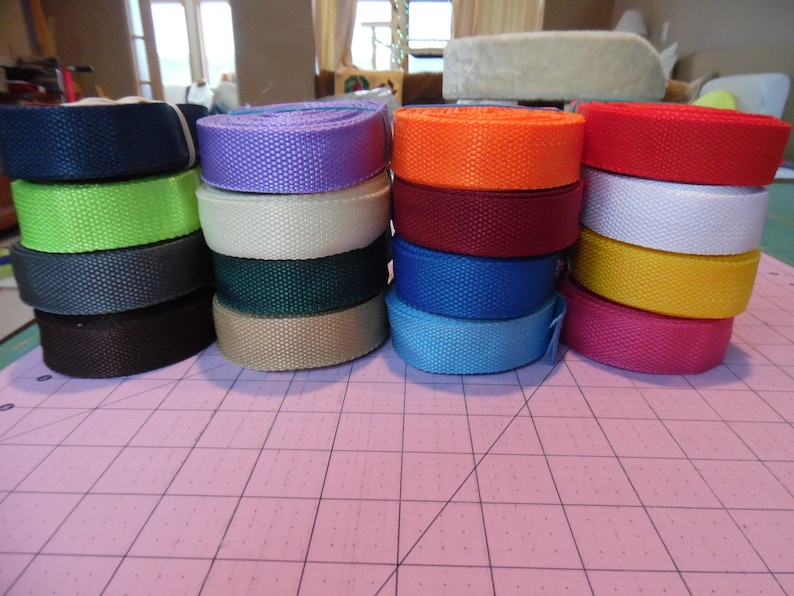 Puede incluir: Una colecci&oacute;n de doce rollos de cinta de nailon tejida dispuestos en cuatro pilas verticales. Los rollos presentan una variedad de colores s&oacute;lidos, incluyendo negro, verde lima, gris oscuro, marr&oacute;n, lavanda, crema, verde oscuro, beige, naranja, granate, azul, rojo, blanco, amarillo y rosa.