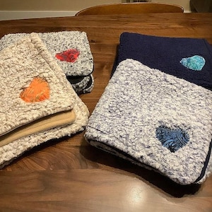May include: Several folded sherpa blankets in various colors, including cream, gray, and navy blue, are displayed on a wooden surface. Each blanket features a heart-shaped patch in contrasting colors, such as orange, red, and teal.