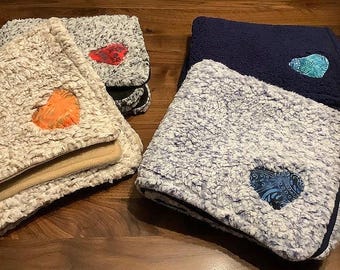 Puede incluir: Varias mantas de sherpa dobladas en varios colores, incluyendo crema, gris y azul marino, se muestran sobre una superficie de madera. Cada manta presenta un parche en forma de corazón en colores contrastantes, como naranja, rojo y turquesa.