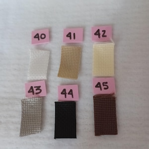 Puede incluir: Seis muestras de tela rectangulares dispuestas en dos filas, etiquetadas con tarjetas de papel rosa numeradas del 40 al 45. Las muestras presentan varios colores s&oacute;lidos, incluyendo blanco, beige, crema, gris claro, negro y marr&oacute;n oscuro.