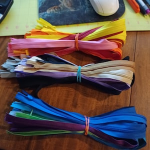 May include: Bundles of colorful zippers in various shades, including pink, yellow, blue, and purple, are secured with rubber bands. The zippers are arranged on a wooden surface, showcasing a variety of colors for sewing and crafting projects.