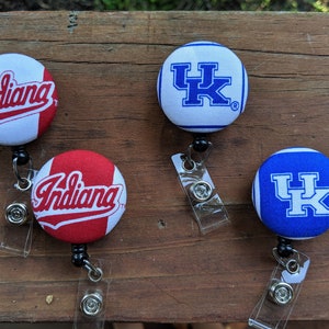 May include: Four fabric badge holders with retractable reels. Two holders feature a white background with red stripes and the word "Indiana" in red. The other two holders feature a white background with blue stripes and the letters "UK" in blue.