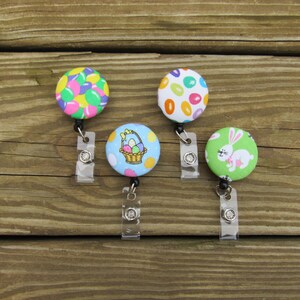 May include: Four Easter-themed fabric badge reels with clear plastic retractable cords. The reels feature a basket of Easter eggs, a bunny rabbit, and two different patterns of colorful jelly beans.