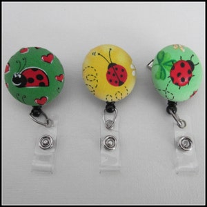 May include: Three colorful ladybug themed retractable badge holders with clear plastic straps. The badge holders are round and feature different ladybug designs on a green, yellow, and green background.