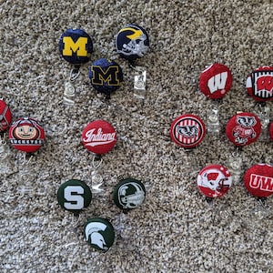 May include: A collection of 14 different college team themed retractable badge holders. The badge holders are round and feature various team logos and mascots, including the University of Michigan, Ohio State, Wisconsin, Indiana, and the University of Washington. The badge holders are all red, white, and blue.